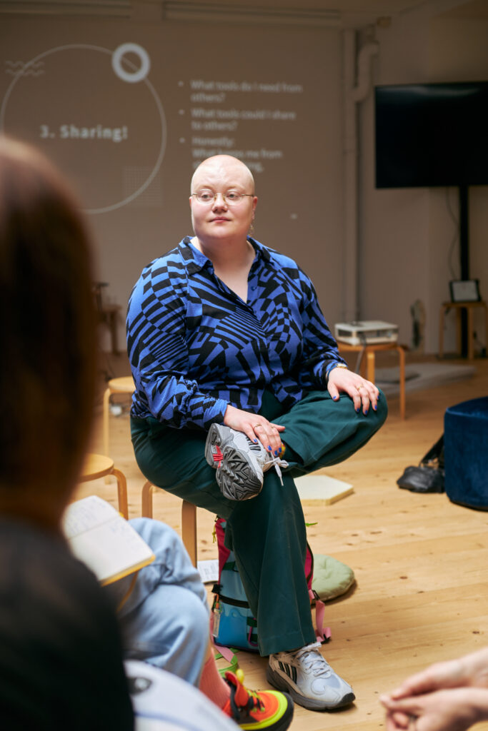 A photo of Isa Hukka facilitating a training. They are sitting on a chair listening, and behind them is a powerpoint presentation.